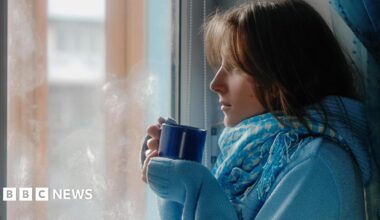 Woman wearing a blue jumper and scarf stands looking out of a window with a warm drink in a mug with steam coming from it in her hand.