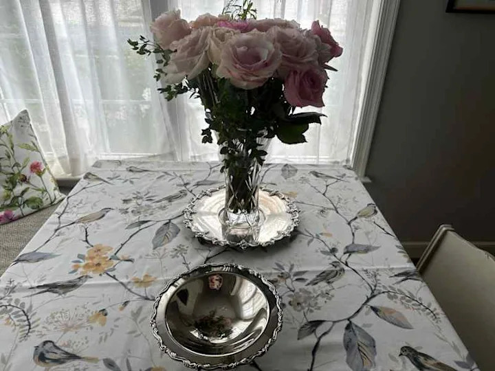 A vase of pink roses on a floral tablecloth with silver serving dishes.