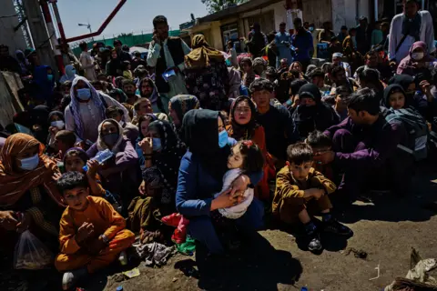 Los Angeles Times via Getty Images Women and children crouch in the sweltering heat at a Taliban-controlled checkpoint near Abbey Gate, an entrance to the Kabul airport on Aug. 25, 2021. They wait to make their way towards the British military-controlled entrance of the airport