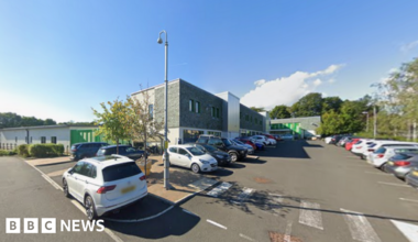 A street view image of Ferndene which is a modern, rectangular building with grey bricks on the front of the building. There are cars parked in front. Large trees can be seen behind the building.
