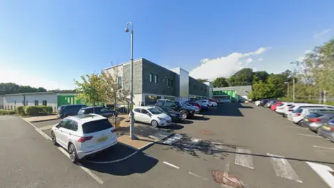 Google A street view image of Ferndene which is a modern, rectangular building with grey bricks on the front of the building. There are cars parked in front. Large trees can be seen behind the building.