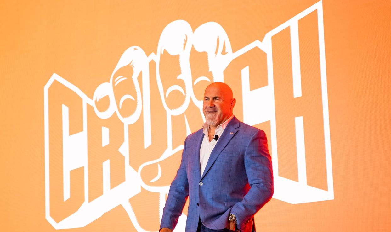 Crunch Fitness CEO Jim Rowley stands in a blue suit in front of the Crunch logo