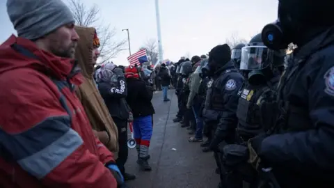Reuters Demonstrators stand in front of members of U.S. Customs and Border Protection (CBP) and other law enforcement officials, near the Bishop Henry Whipple Federal Building. One man is wearing a red jacket another is wearing a brown jacket opposite a line officials wearing all black and black helmets
