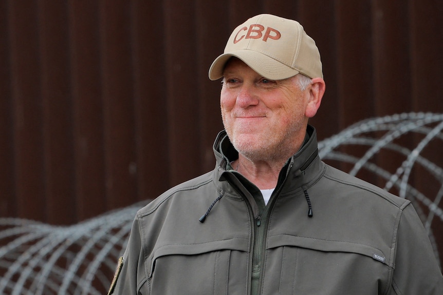 A man wearing a beige baseball cap stands in front of coils of barbed wire and smiles
