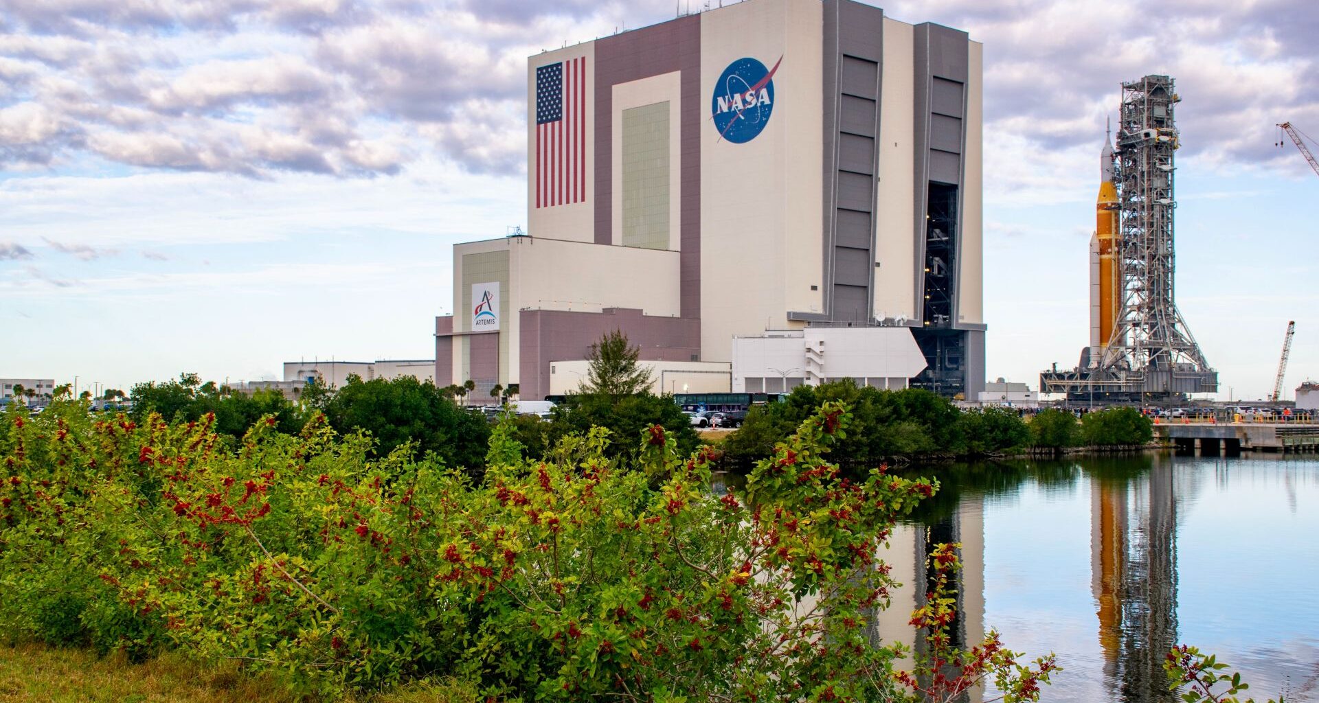 An orange rocket sits on a mobile scaffold as it rolls out of a large white building with the American flag and NASA meatball logo on the side.