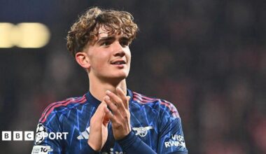 Arsenal midfielder Max Dowman applauds as he celebrates the team's victory in the Champions League match against Slavia Prague. He is wearing a blue patterned Arsenal shirt, with three red stripes as trim.