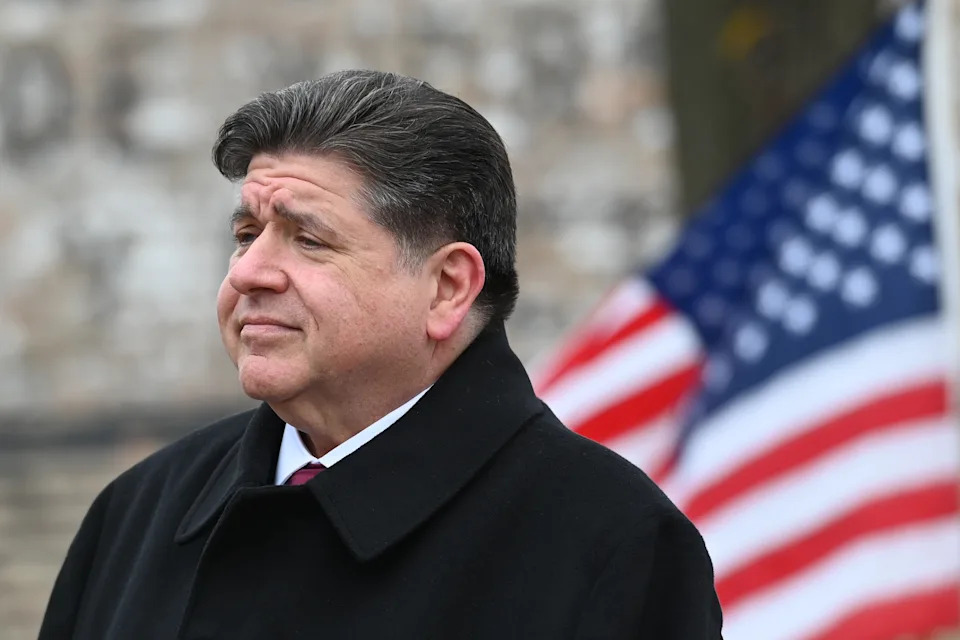 Person in a suit stands outdoors with an American flag in the background