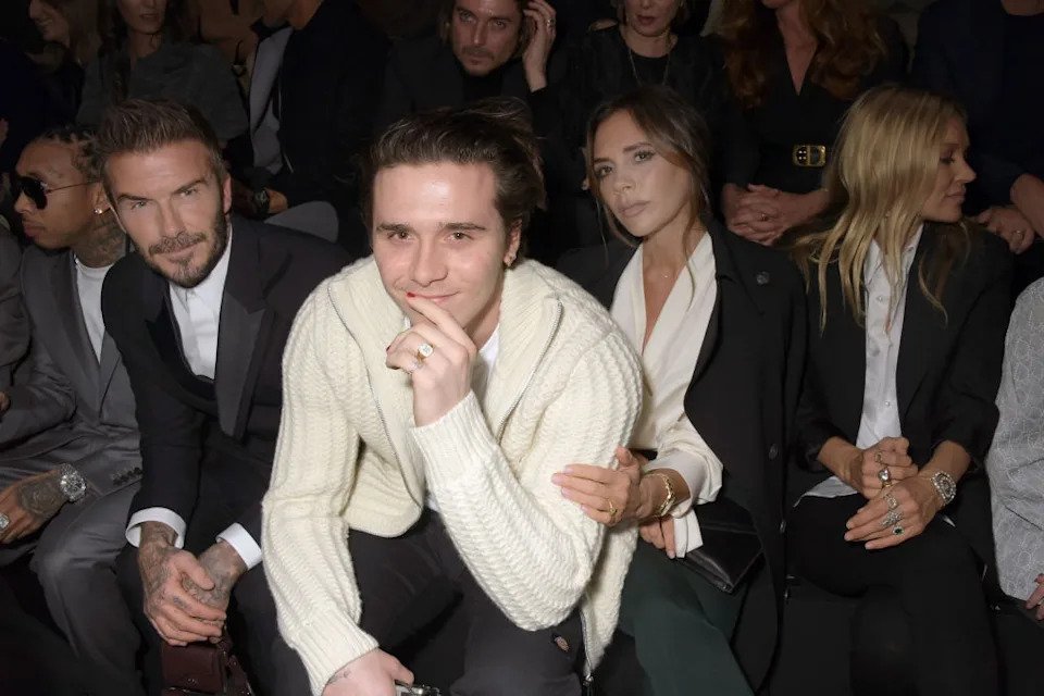 A group of people, including a family, sits in a front row at a fashion event. The son wears a knitted sweater; the parents are dressed in chic outfits