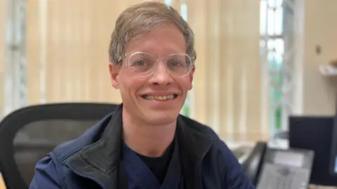 Dr Chris Jacobs smiles at the camera. He is wearing glasses, a blue gilet and medical scrubs top. He sits in a computer chair.