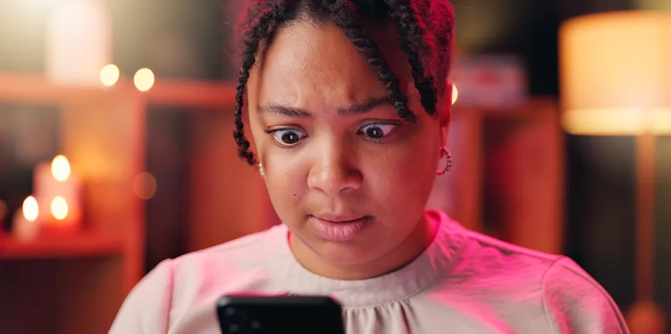 A person looks surprised while reading something on their phone, seated indoors with decorative lights in the background
