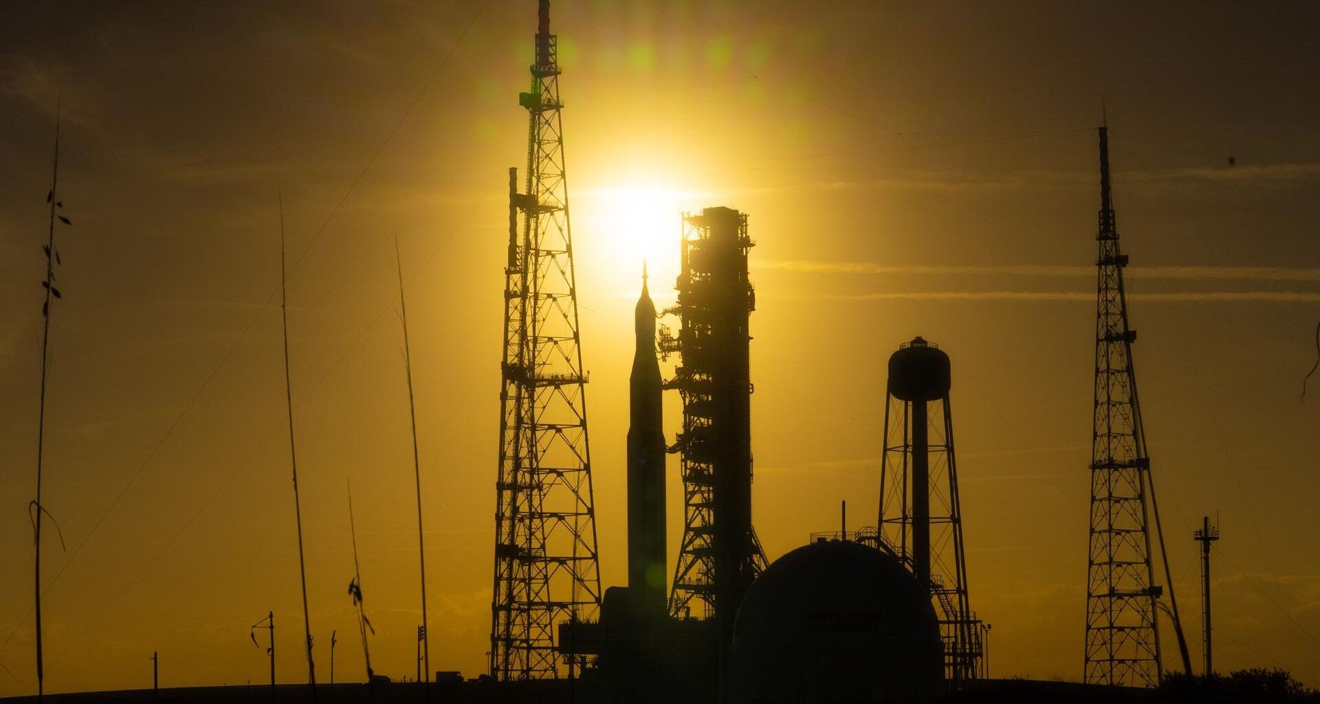 A silhouette of a rocket surrounded by scaffolding is seen in a dim yellow sunset