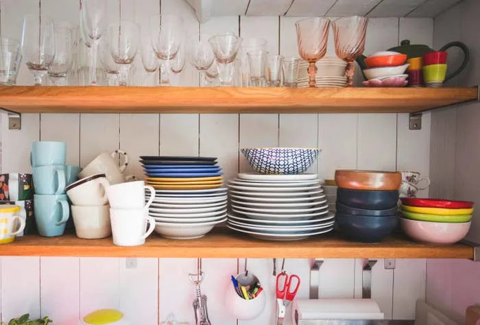 Open kitchen shelves with various stacked plates, bowls, mugs, and glasses. Colorful dishes add a lively touch to the organized display