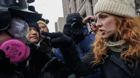 Getty Images A woman with red hair and wearing a white cable-knit beanie hat points a gloved finger at a man dressed in googles, a hardhat and a pink respirator mask. She looks angry but not afraid. the action is happening in a crowded group of people, a cameraman is taking photographs behind them.