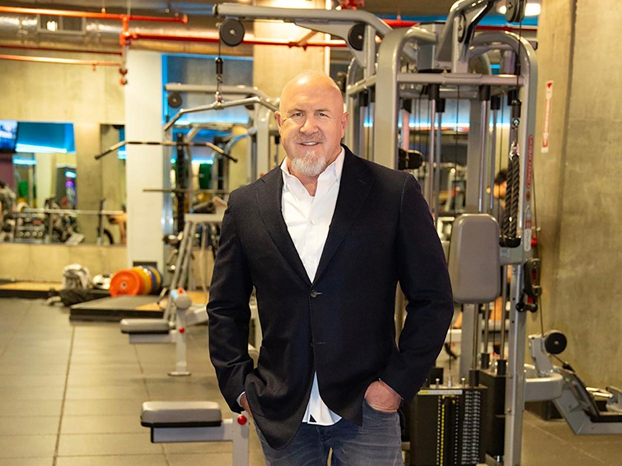 a man in a formal shirt and blazer and jeans smiling at the camera in a gym