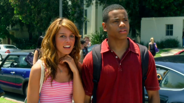 Shenae Grimes as Annie in pink tank top standing next to Tristan Mack Wilds as Dixon with fancy cars behind them in 90210