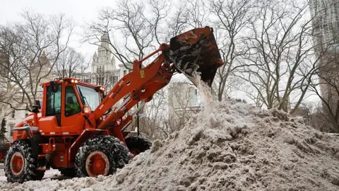 Getty Images An orange plow dumps snow onto a large, grey snow pile in New York City.  City Hall can be seen in the background.
