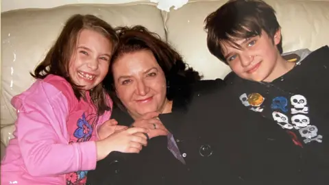 Ava Randle Ava (on the left, in pink, smiling) and Owain (on the right, in a dark hoodie) when they were younger with their mum Keren (in the middle in dark clothes, with long brown hair)