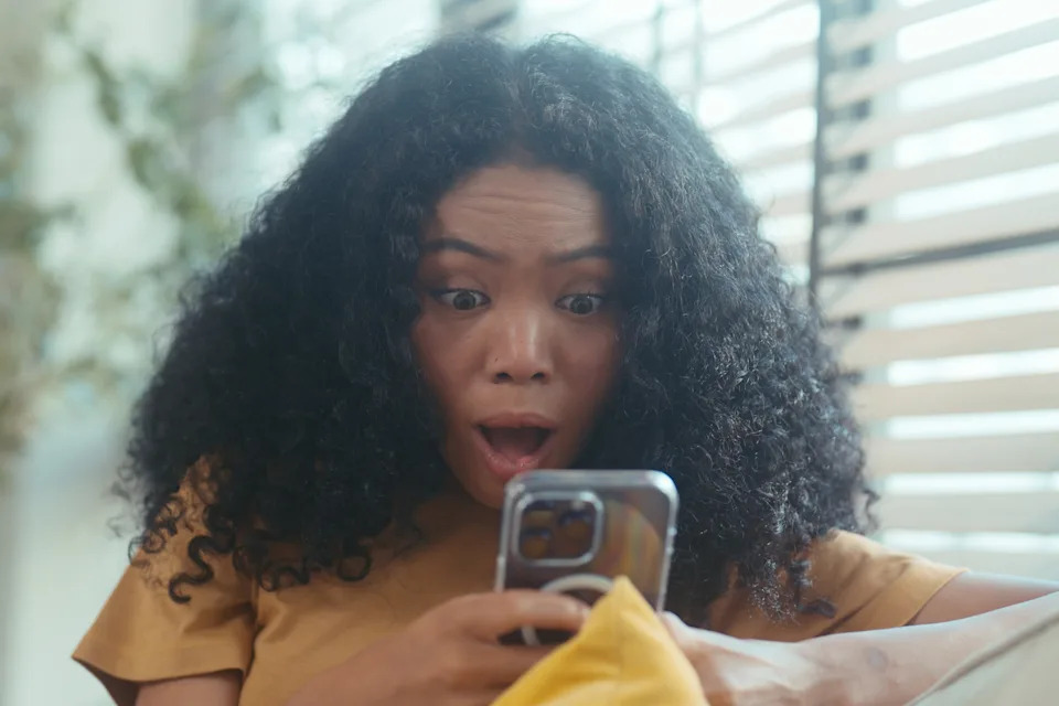 Person with curly hair, wearing a casual outfit, looks surprised while holding a smartphone