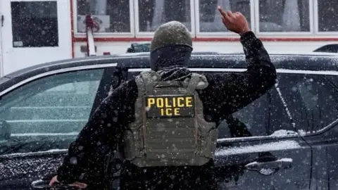 Reuters An Immigration and Customs Enforcement agent raises a finger moments after detaining a man during an immigration raid, he is standing with his back to the camera and is wearing all black apart from a green vest with the words ICE on it in yellow writing 