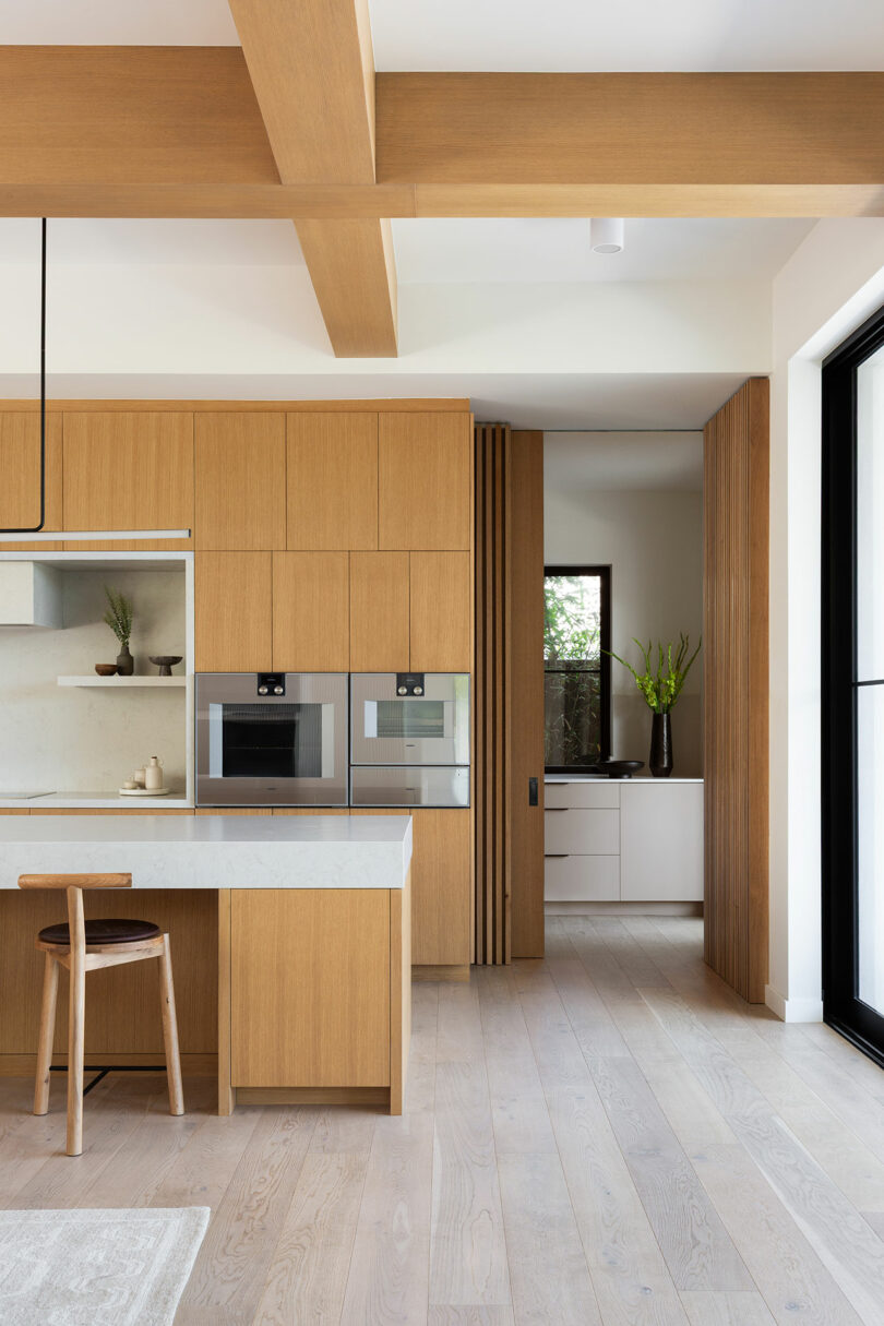 Modern kitchen with light wood cabinets, built-in ovens, a white countertop island with a wooden stool, and large windows providing natural light.