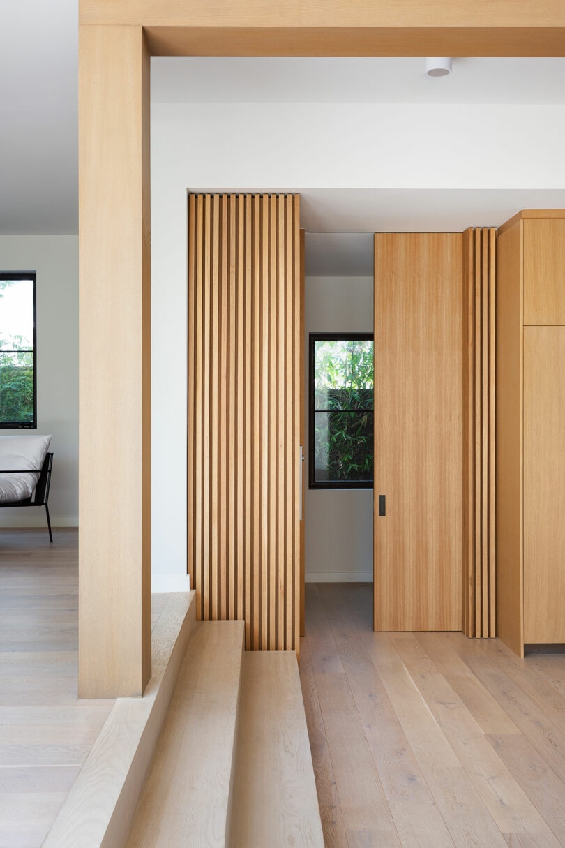 Minimalist interior with light wood finishes, vertical slatted wood panels, split-level flooring, and a window showing greenery outside. A portion of a bed is visible in an adjacent room.