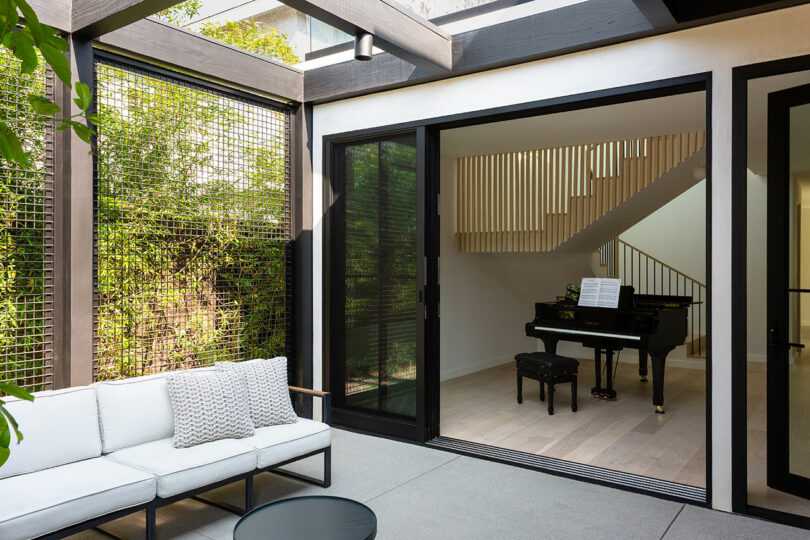 A modern living area with a white outdoor sofa and table opens to an indoor space featuring a black grand piano and staircase with vertical wooden slats.