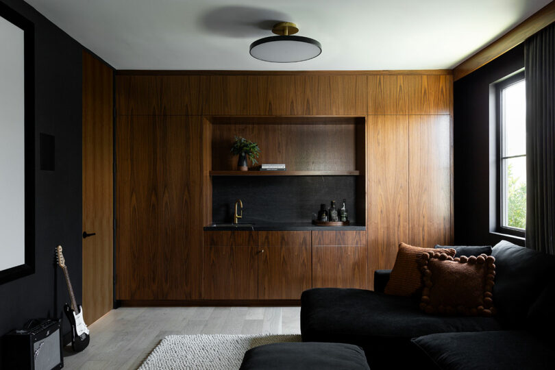 A modern living room with dark wood cabinetry, a built-in sink, shelves with decor, a black sectional sofa, and a window with gray curtains.