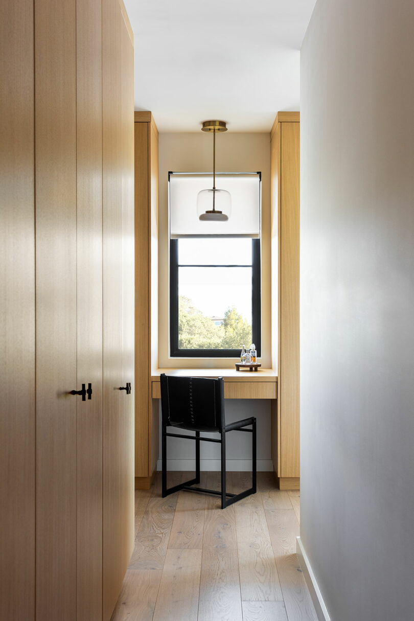 A small home office nook with a wooden desk, black chair, pendant light, and window overlooking greenery, set between light wood cabinets and closet doors.