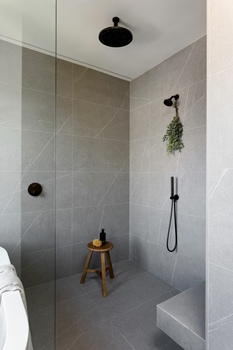 Modern bathroom with gray large-format tiles, walk-in shower, black fixtures, a small wooden stool with a sponge and bottle, and greenery hanging from the wall.