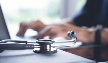 A stethoscope lies on a desk in the foreground, symbolizing health care. In the blurred background, hands type on a laptop, suggesting they are using the Federal Electronic Health Record Management system.