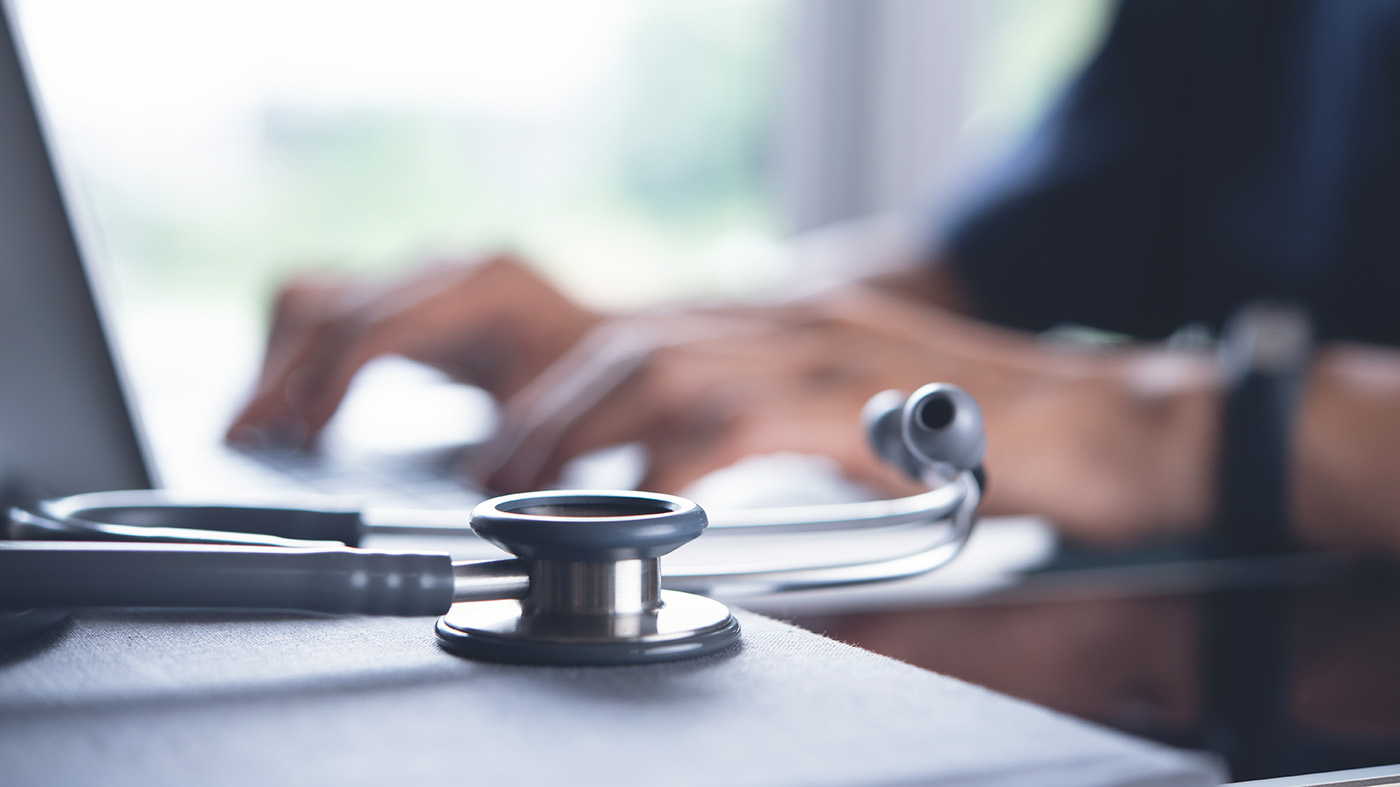 A stethoscope lies on a desk in the foreground, symbolizing health care. In the blurred background, hands type on a laptop, suggesting they are using the Federal Electronic Health Record Management system.