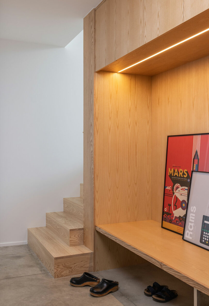 Minimalist entryway with light wood bench and wall, built-in lighting, framed posters, wooden stairs, and shoes on concrete floor.