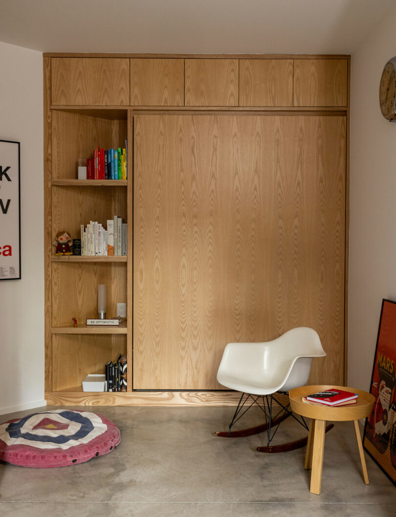 A wooden built-in shelving unit with books and decor next to a white rocking chair, small round table with a red book, and a round rug on a concrete floor.