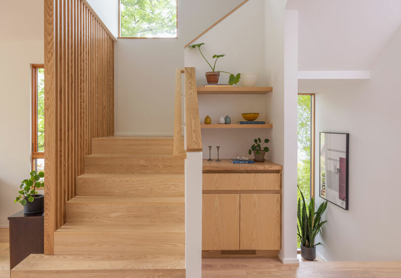 Modern wooden staircase with vertical slat railing beside built-in shelves with plants and decor; large windows provide natural light.