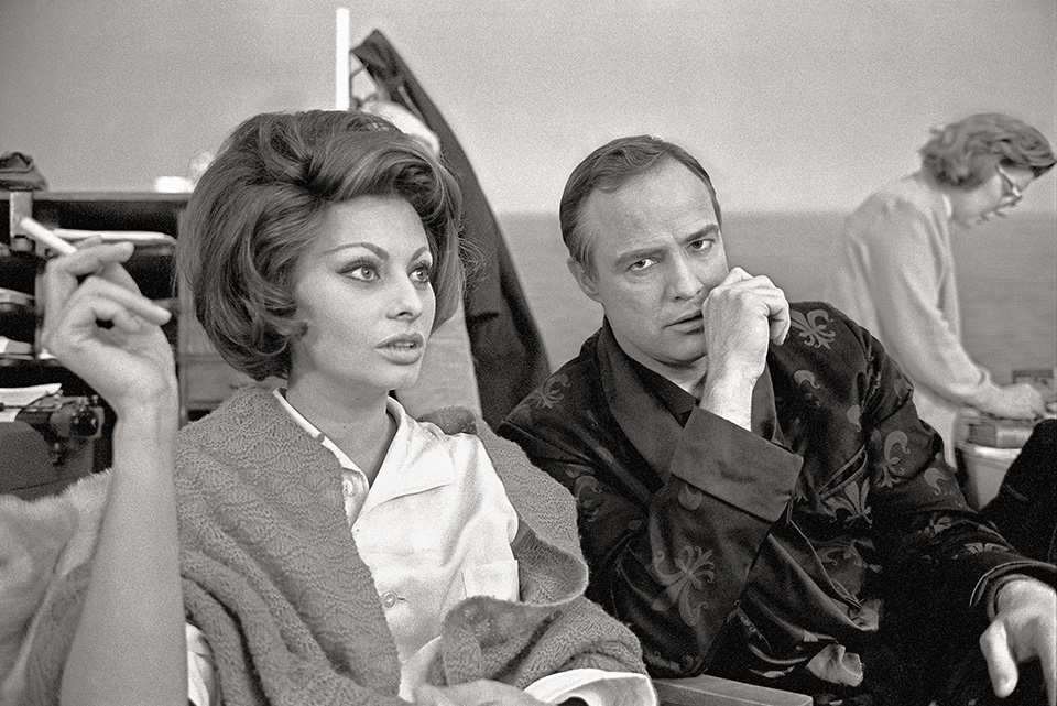 A black-and-white candid shot features Sophia Loren holding a cigarette and looking to the side while sitting next to Marlon Brando.