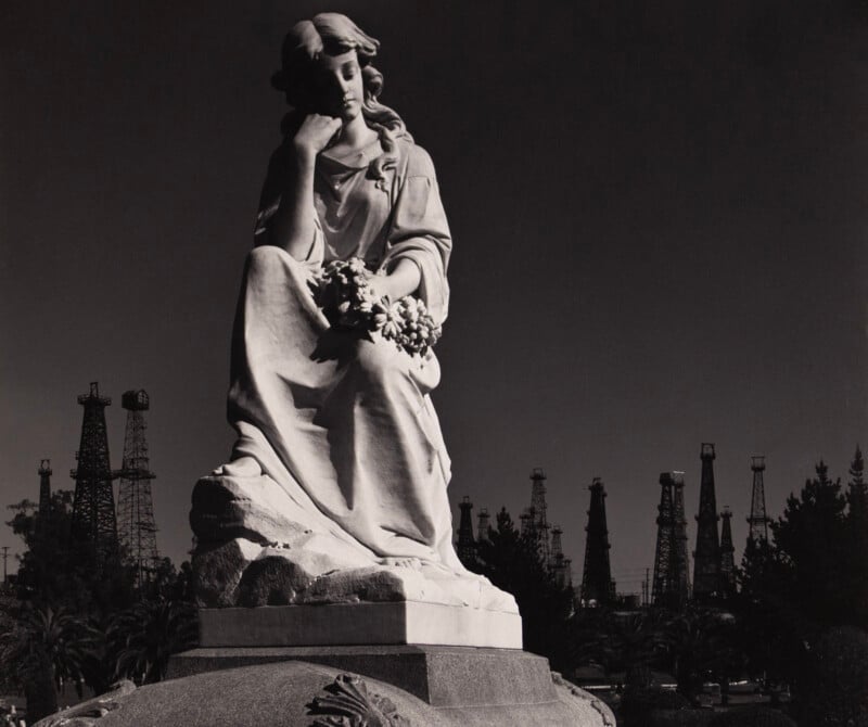 A marble statue of a thoughtful woman holding flowers is set against a dark sky, with several oil derricks visible in the background.
