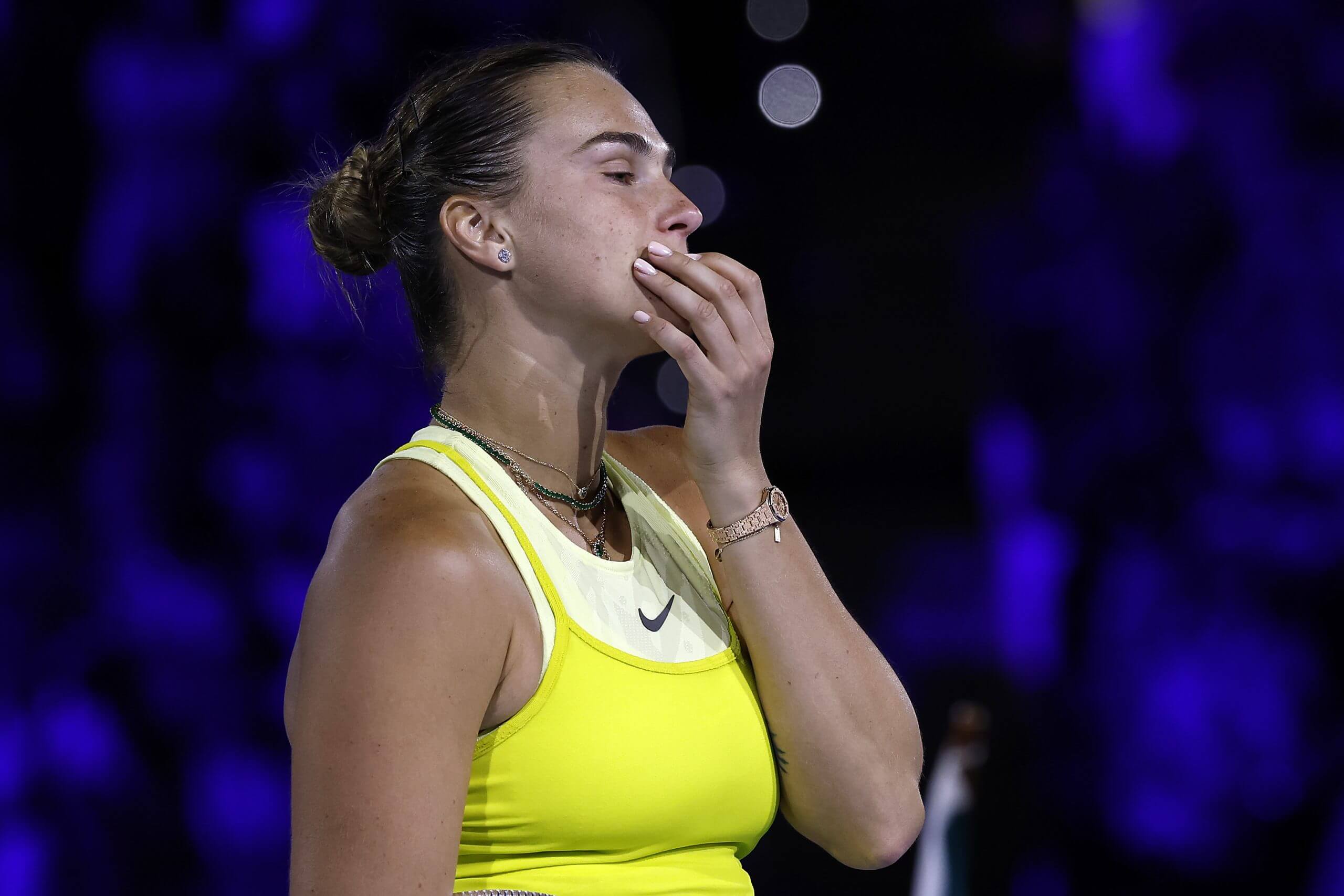 Aryna Sabalenka looks pensive with her left hand raised to her chin as she looks on wearing a yellow tennis outfit.