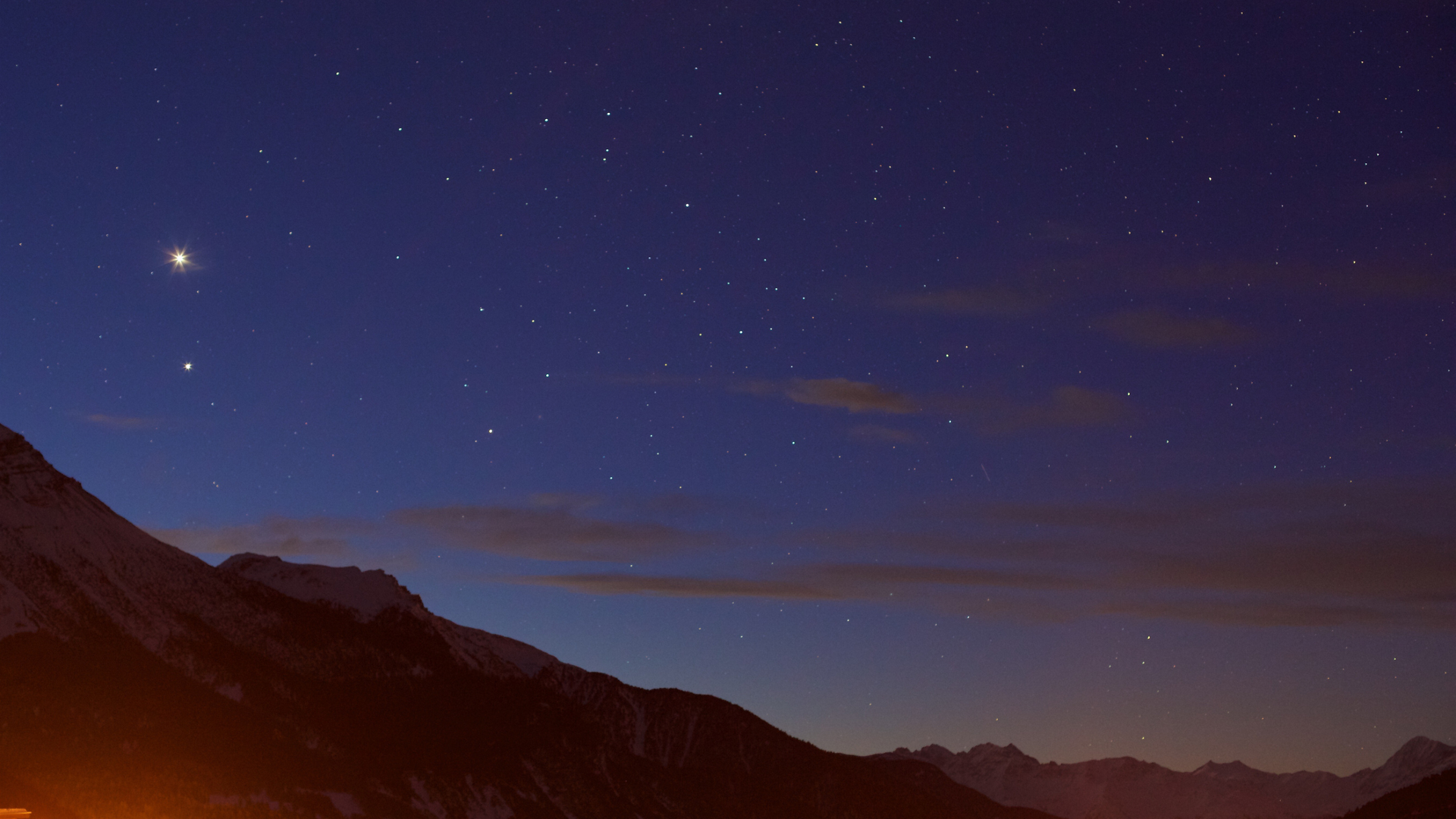 In a royal blue night sky, a bright dot for the planet Jupiter is seen toward the left of the image with dark pink clouds in the sky obscuring some of the stars. Red cliffs are seen in the foreground of the image