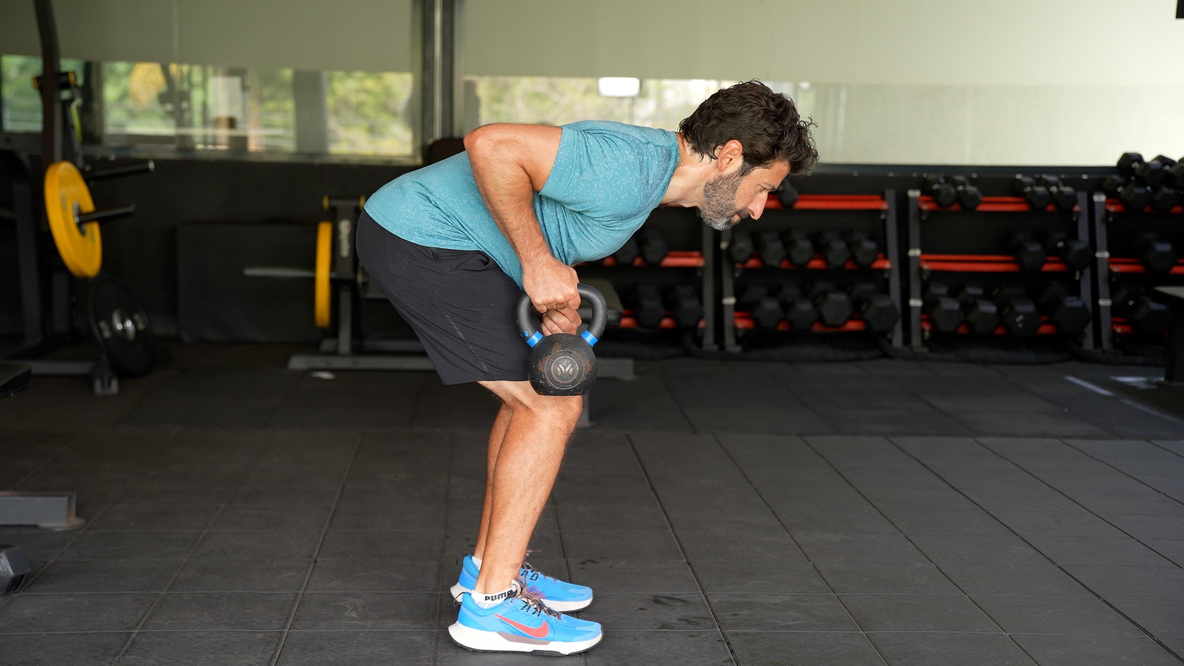 a man with dark hair wearing a blue shirt and black shorts demonstrates a bent-over kettlebell row