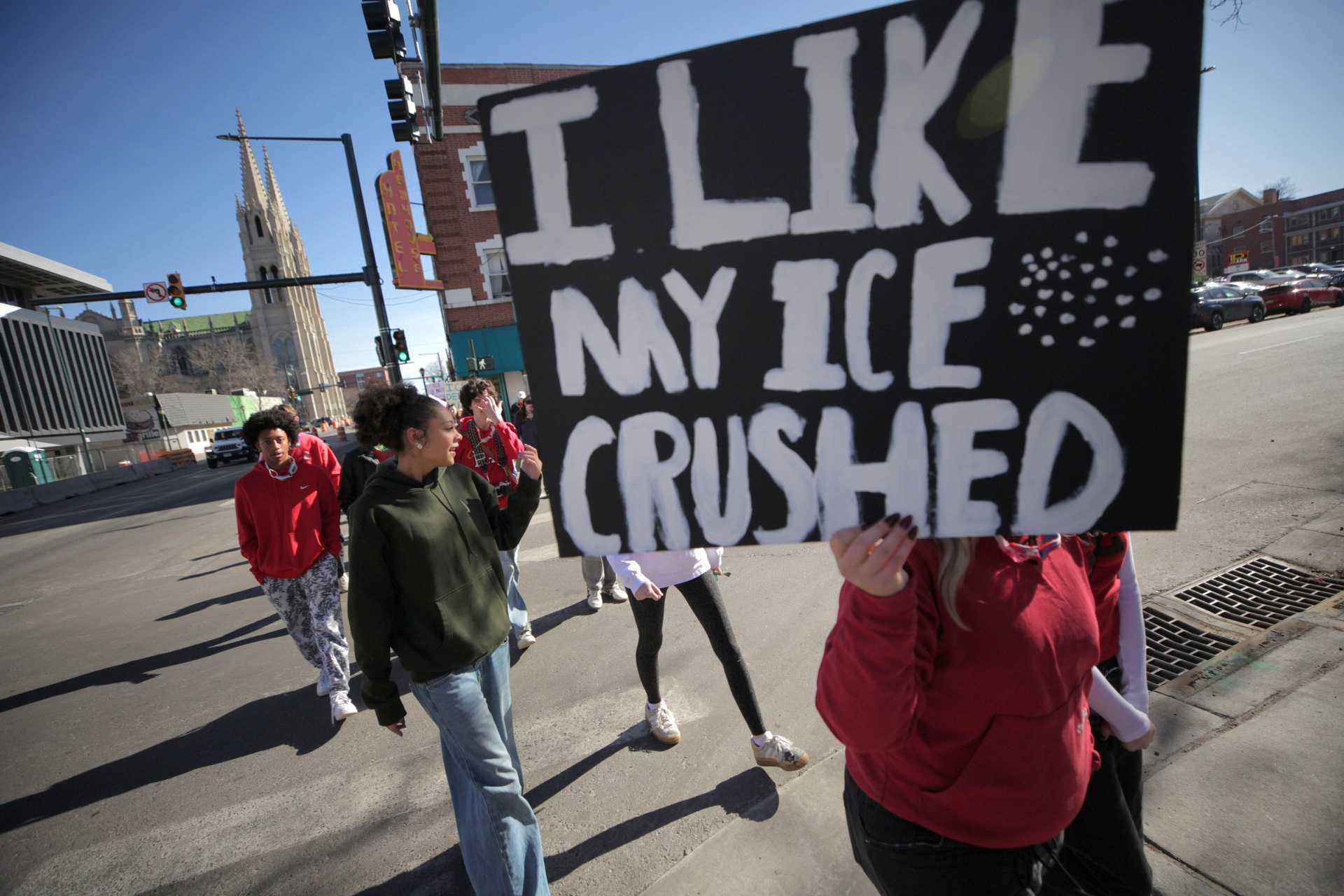 Several high school students, most dressed in red shirts, cross a street. Most of the photo is covered by a large black sign that one student is holding up. The sign reads in hand-painted white letters: "I like my ice crushed"