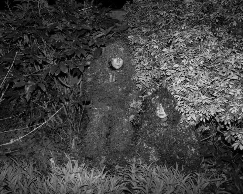 Two people in camouflage suits, blending into dense bushes and foliage at night. Both have serious expressions, with one standing and the other crouching among the plants. The image is in black and white.