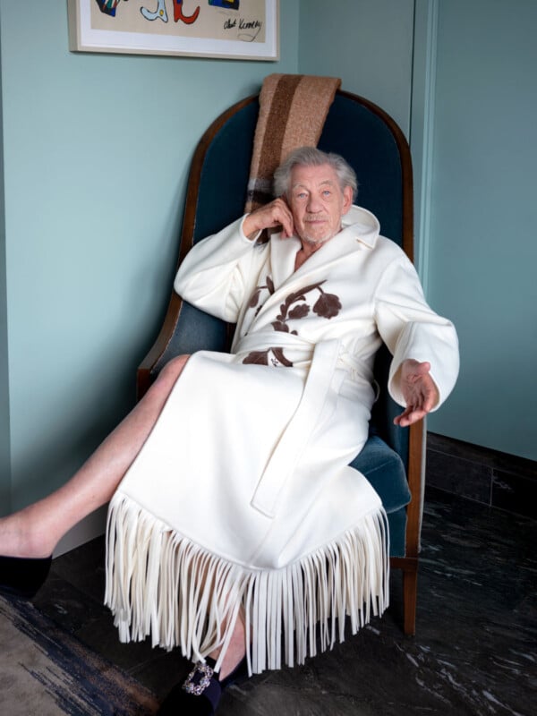 An older man sits on a high-backed chair, wearing a white robe with brown designs and fringes. He has short gray hair, rests one arm on the chair’s side, and extends the other, with legs crossed and one slipper visible.