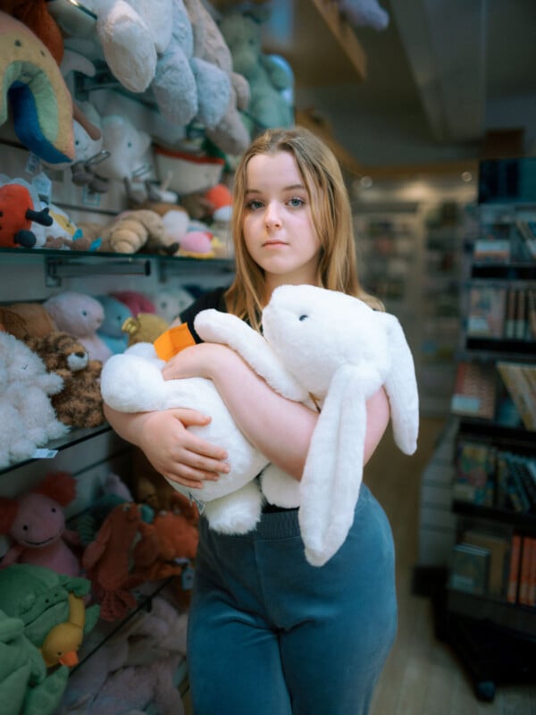 A young person with long blonde hair stands in a toy store, holding a large white plush rabbit. Shelves filled with various stuffed animals and toys are in the background. The person looks calmly at the camera.