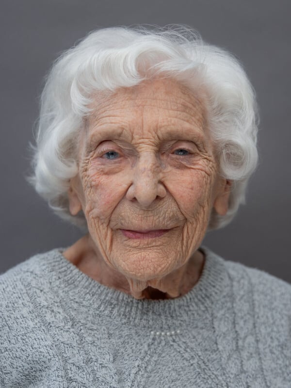 An elderly woman with white hair, wearing a grey knitted sweater, smiles gently while looking at the camera against a plain grey background.