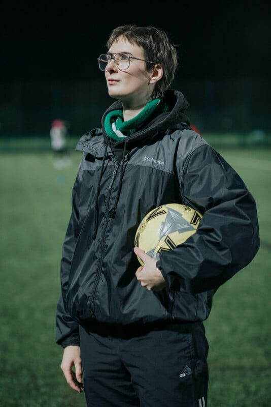 A person wearing glasses and a black jacket stands on a soccer field at night, holding a yellow soccer ball under one arm and looking upwards. The background is blurred with faint figures and lights.