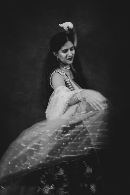 A woman in traditional attire dances gracefully, her sheer sari and long hair flowing as she moves. The image is black and white, capturing a sense of motion and elegance against a dark background.