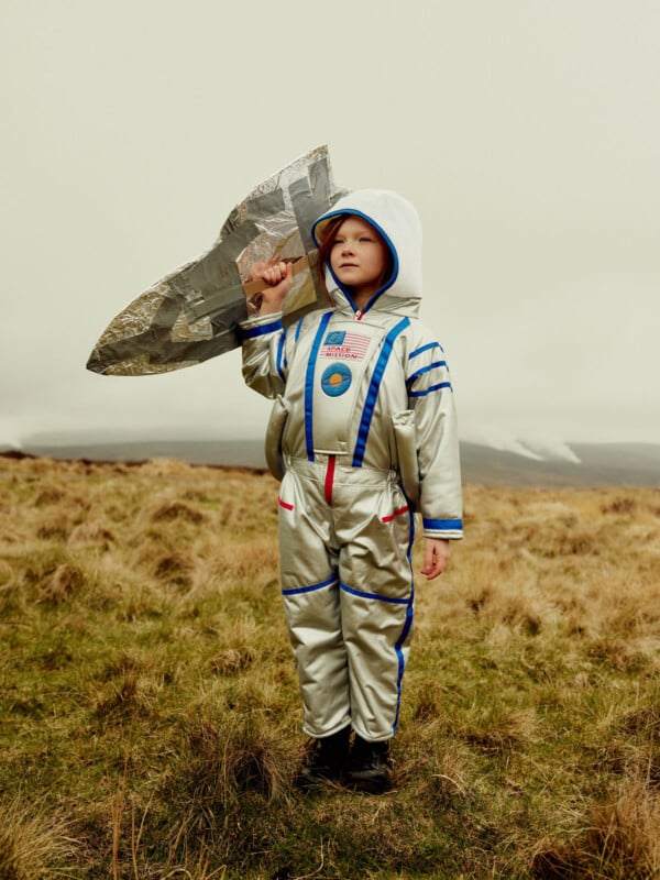 A child wearing a silver astronaut costume and hood stands in a grassy field, holding a homemade foil rocket over their shoulder, with an overcast sky in the background.