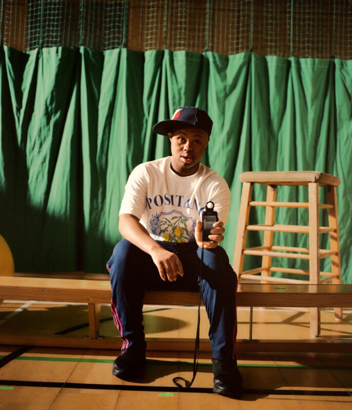 A person in a white graphic t-shirt, navy pants, and a sideways cap sits on a bench in a gym, holding a device. A wooden stool and green curtain are in the background. The scene is warmly lit.