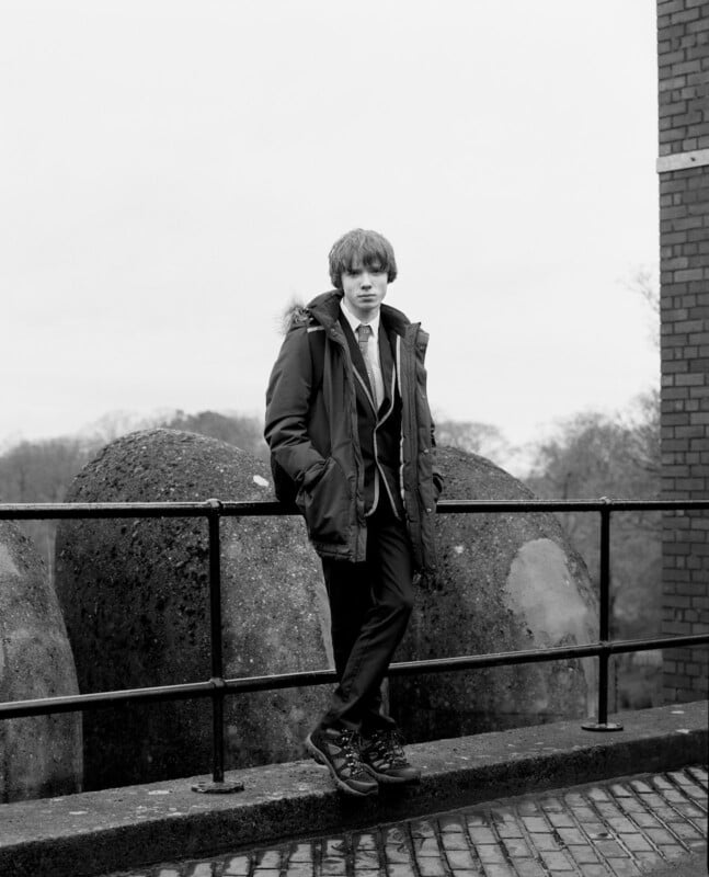 A young person in a winter coat and suit stands with hands in pockets, leaning against a rail outdoors. Large stone shapes and a brick building are in the background. The image is black and white.