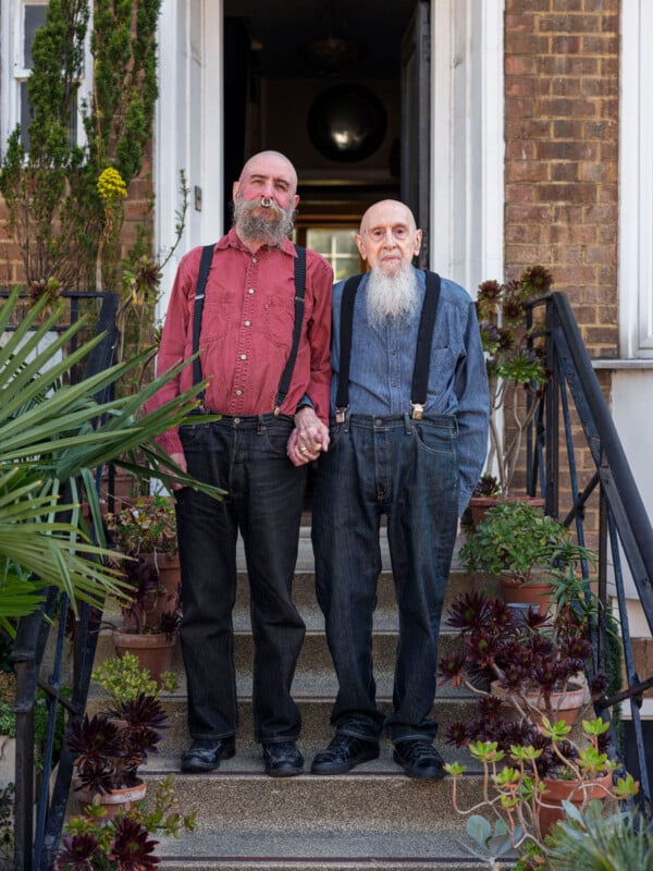 Two older men with beards stand holding hands on a front porch surrounded by potted plants, wearing suspenders and collared shirts, looking at the camera.